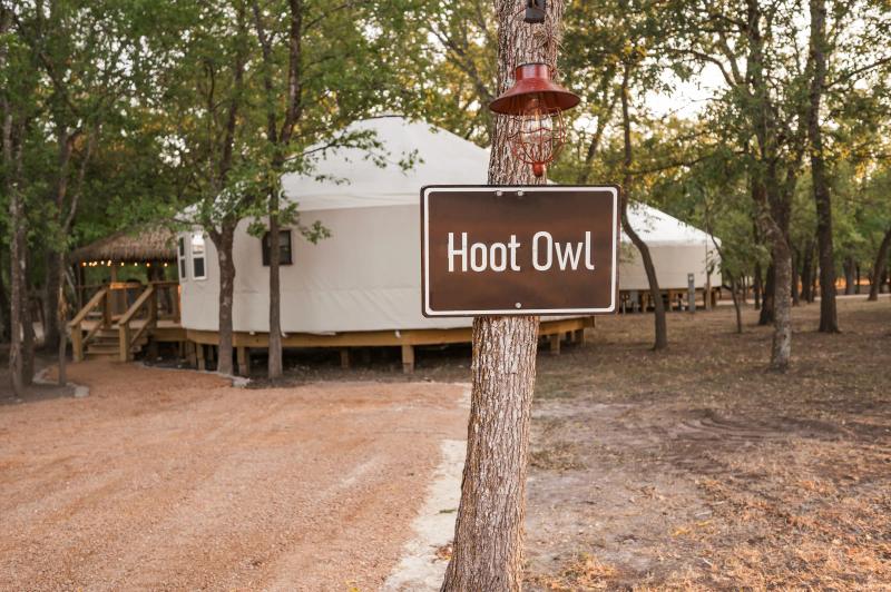 Hoot Owl Yurt — entrance sign beneath live oaks with the canvas yurt beyond.