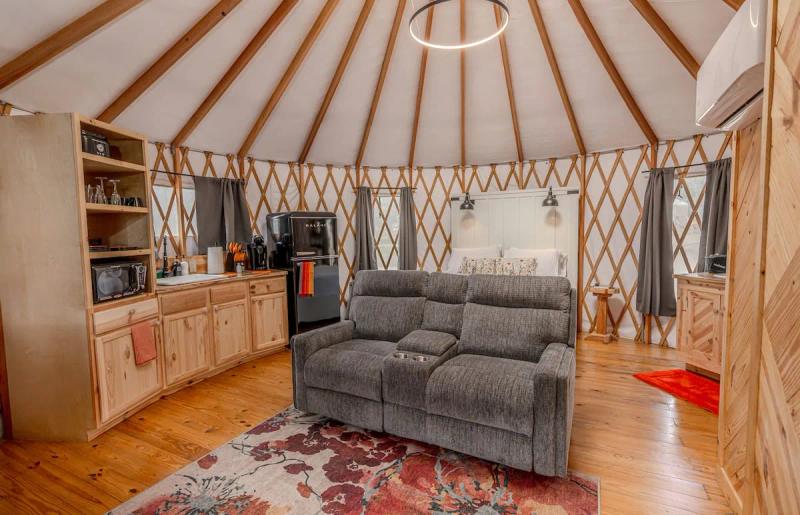 King Fisher Yurt — living area with loveseat and wood kitchenette beneath circular roof beams.