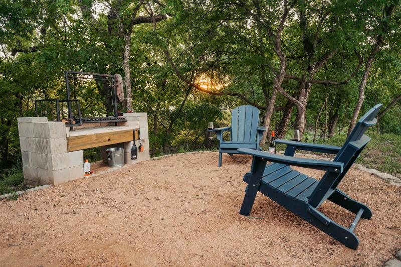 Love Bird Yurt — stone grill and two navy Adirondack chairs with the sun setting through the oaks.