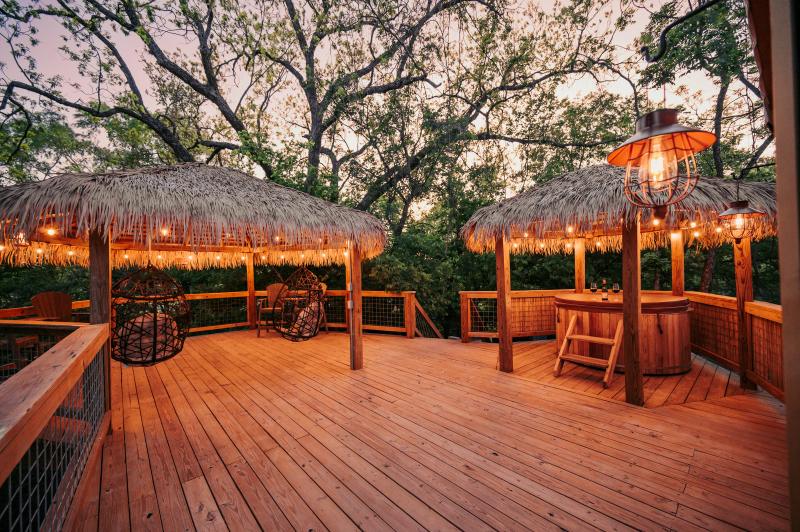 Love Bird Yurt — two thatched gazebo roofs and the hot tub glowing under bulb lights at twilight.