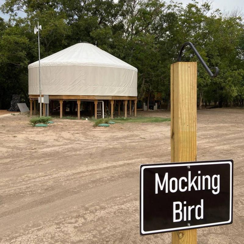 Mockingbird Yurt — hand-lettered Mocking Bird sign at the drive with the yurt on its cedar platform behind.