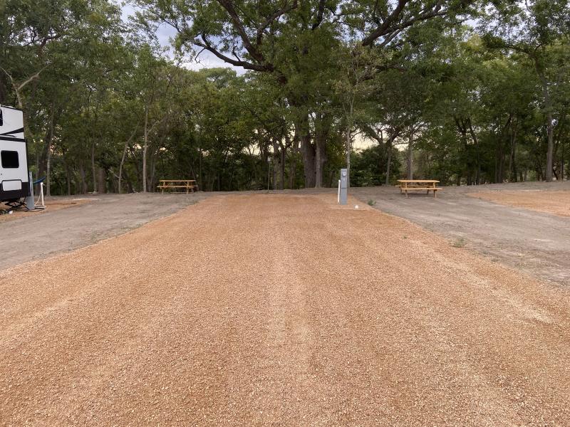 River Yurt Village RV Park — wide gravel RV pads and picnic tables spread out beneath the pecan canopy, a travel trailer parked at the far left.